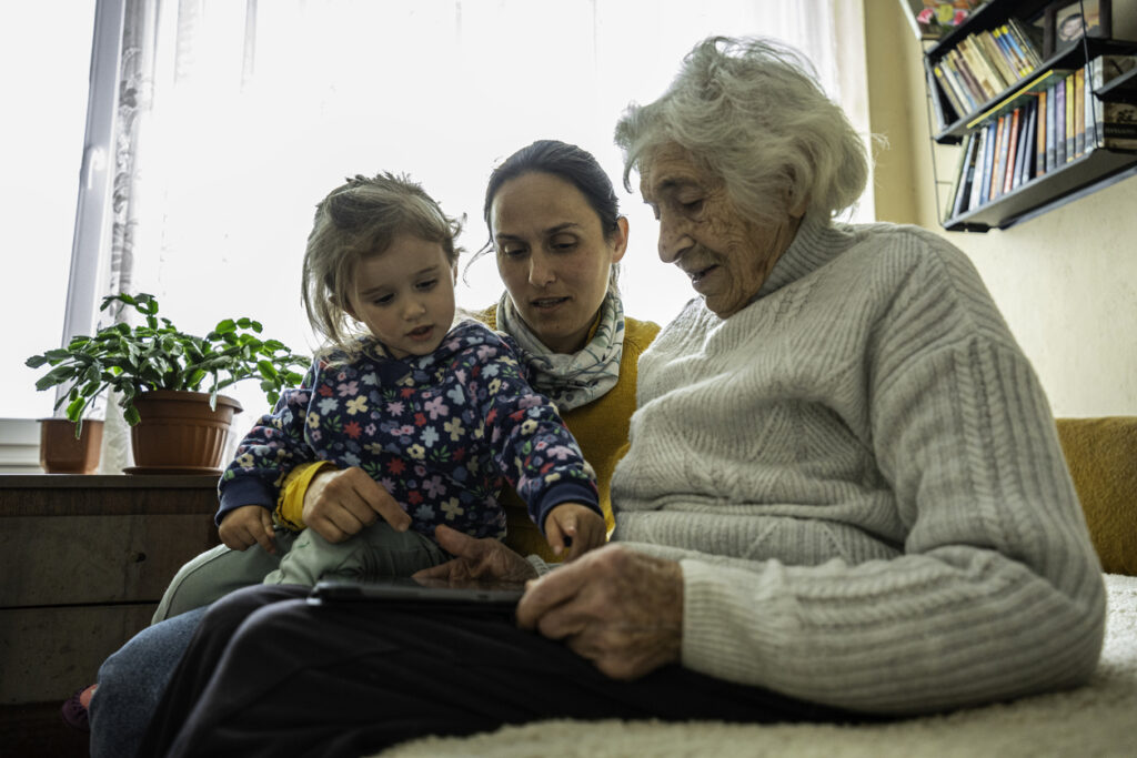 A middle-aged woman sits between a young child and an elderly woman, representing the sandwich generation experience of caring for both children and aging parents.