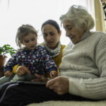 A middle-aged woman sits between a young child and an elderly woman, representing the sandwich generation experience of caring for both children and aging parents.