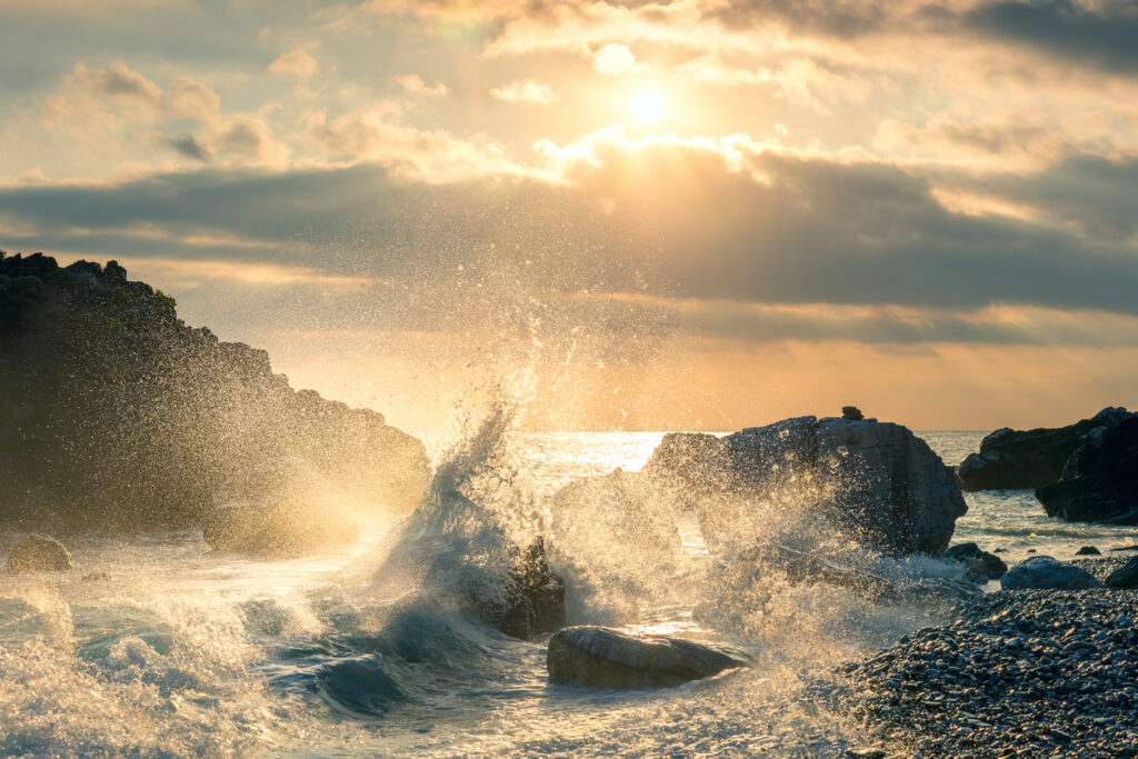 Large ocean wave crashing against coastal rocks with golden sunlight breaking through dramatic storm clouds