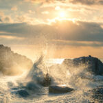 Large ocean wave crashing against coastal rocks with golden sunlight breaking through dramatic storm clouds