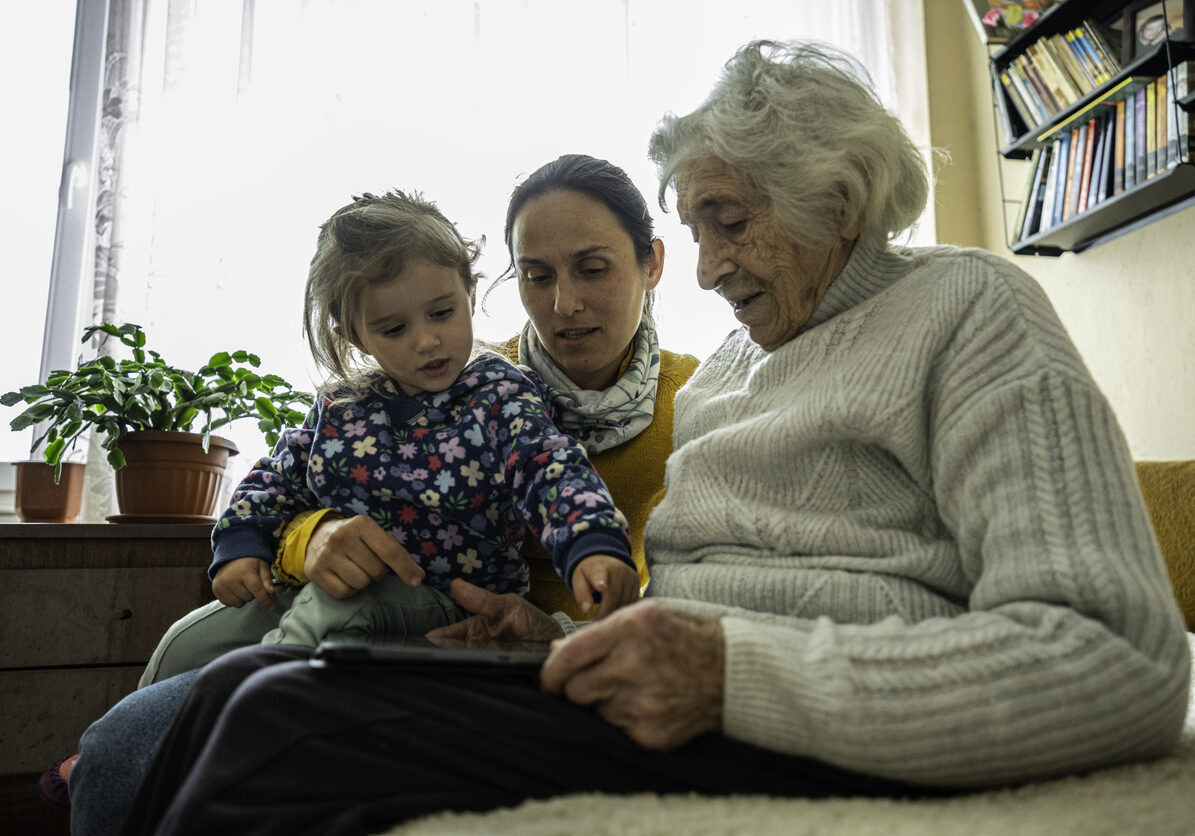 A middle-aged woman sits between a young child and an elderly woman, representing the sandwich generation experience of caring for both children and aging parents.