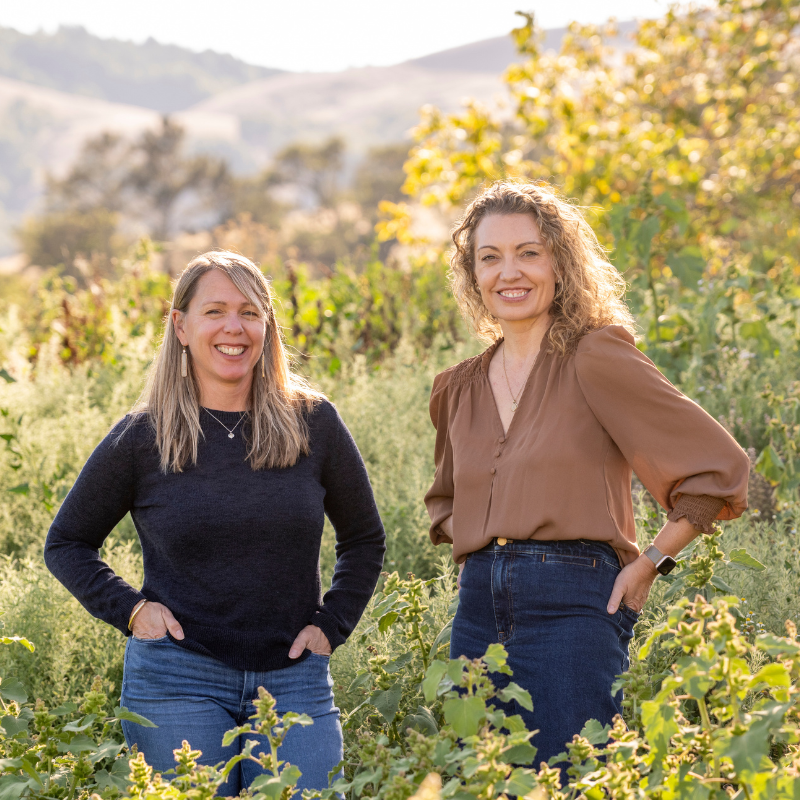 Jamie Katoff and Victoria Whisman, licensed therapists at A Place to Exhale, smiling outdoors in a sunlit vineyard setting