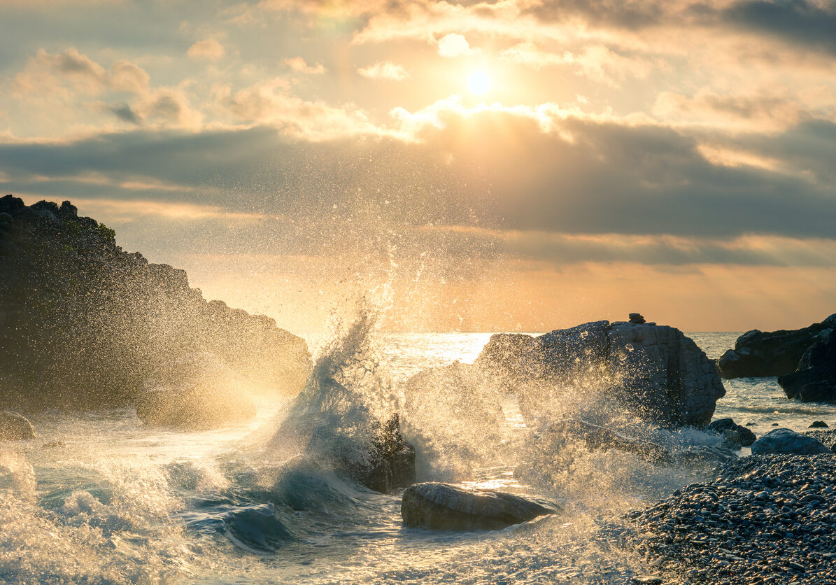 Large ocean wave crashing against coastal rocks with golden sunlight breaking through dramatic storm clouds